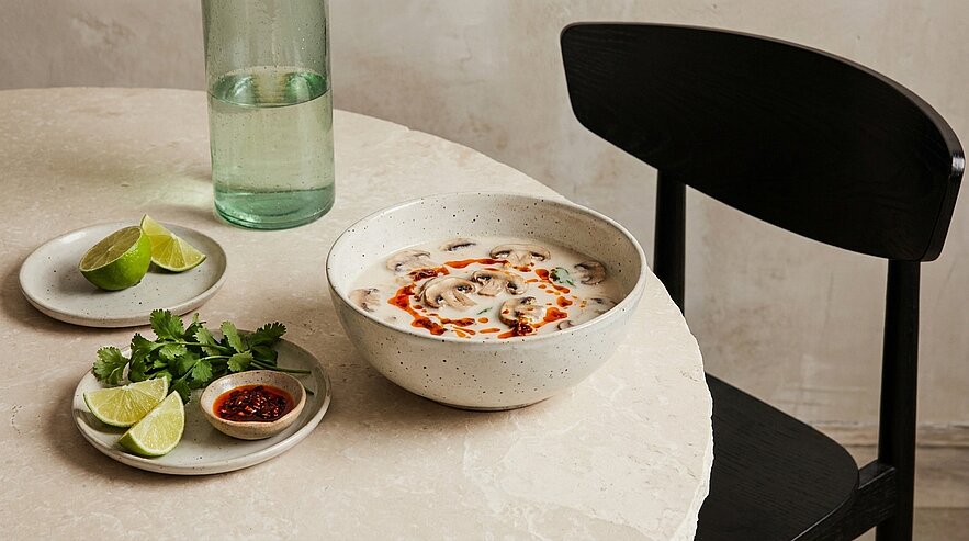 Schale mit cremiger Kokos-Pilzsuppe, garniert mit Chiliöl; daneben kleine Teller mit Limettenstücken und frischem Koriander.