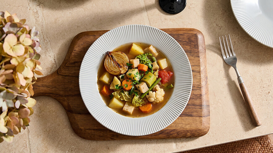 Eine Schüssel mit bunter Gemüsesuppe liegt auf einem Holzbrett. Die Suppe enthält Kartoffeln, Karotten, Blumenkohl und verschiedene grüne Gemüsesorten. Sie wird von einem weißen Rand umgeben und ist in der Mitte der Bildkomposition platziert.