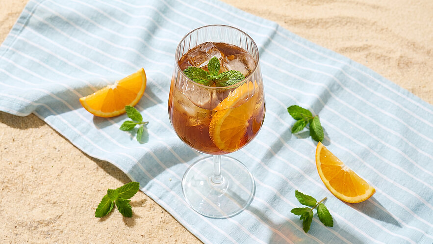 Ein Glast mit einem erfrischenden Getränk, gefüllt mit Eis, Orangenscheiben und einem Minzblatt. Das Glas steht auf einem gestreiften Tuch, umgeben von weiteren Orangenscheiben und frischen Minzblättern, vor einem sandigen Hintergrund.