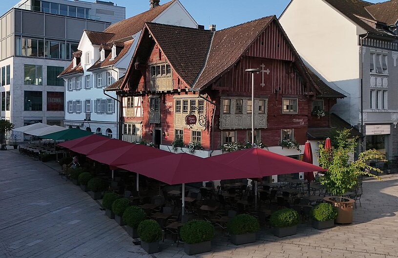Ein historisches Gebäude mit roten Holzverkleidungen und Fensterläden befindet sich zwischen modernen Bauten. Vor dem Haus gibt es einen Außenbereich mit Tischen und roten Sonnenschirmen. Der Platz ist tagsüber sonnig und einladend gestaltet.