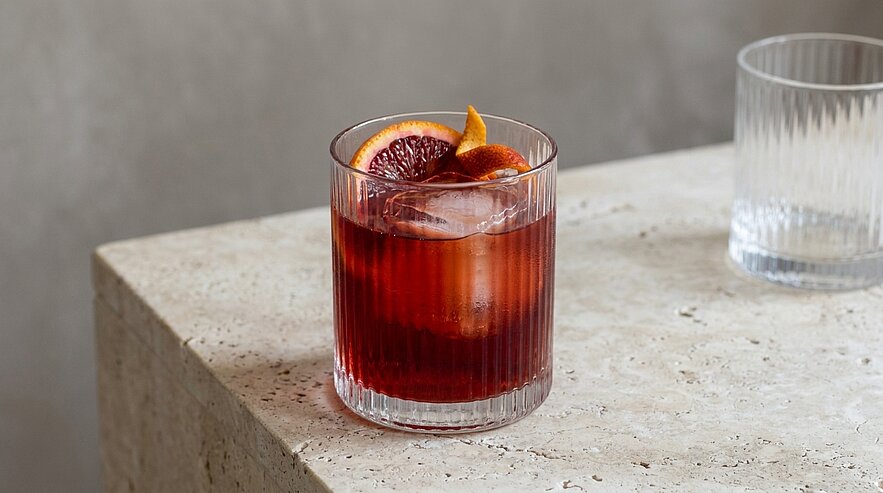 Roter Cocktail in einem kurzen Tumbler mit Eiswürfeln und einer Blutorangenscheibe als Garnitur auf hellem Steintisch.
