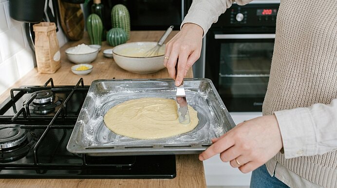 Person verstreicht mit einem Teigschaber runden Teig auf einem bemehlten Backblech in einer Küche.