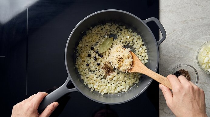 Zwei Hände rühren mit einem Holzlöffel fein gewürfelte Zwiebeln und Gewürze (Lorbeerblatt, Wacholderbeeren) in einem Topf auf dem Herd.
