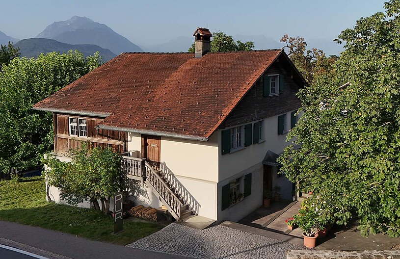 Ein zweigeschossiges, traditionelles Haus mit einem roten Dach und Holzverkleidung an einer Seite. Eine Treppe führt zur Eingangstür, umgeben von grünen Bäumen und Sträuchern. Im Hintergrund sind sanfte Berge zu sehen, während die Sonne den Himmel erhellt.