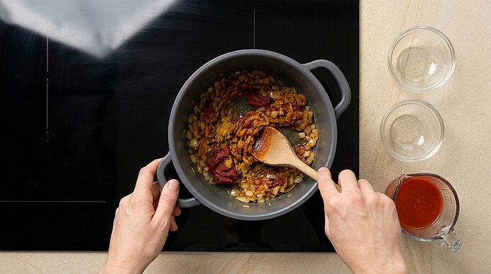 Hände rühren mit einem Holzlöffel eine Tomaten‑Zwiebel‑Gemüse‑Mischung in einem Kochtopf auf dem Herd.