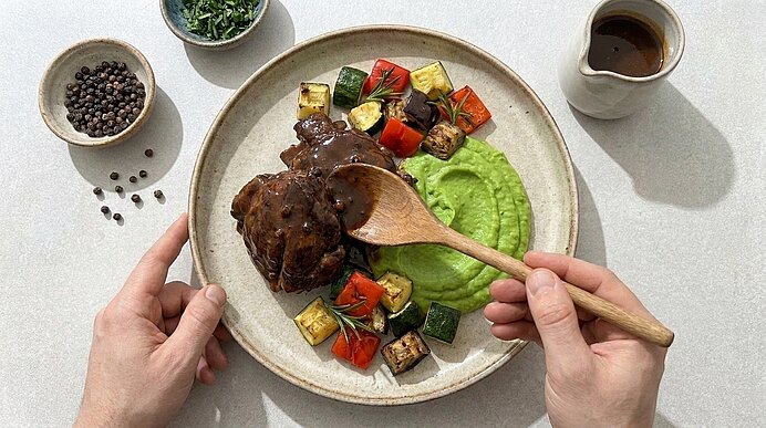 Zwei Hände servieren einen Teller mit geschmortem Fleisch in dunkler Sauce, grünem Püree und gerösteten Gemüsewürfeln.