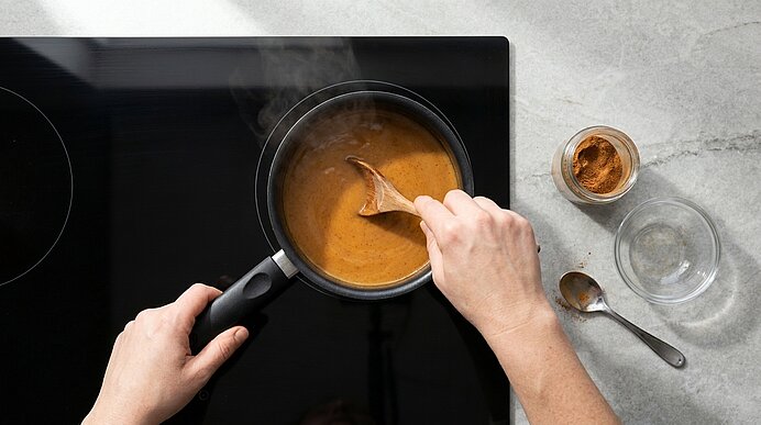 Hände rühren mit einem Holzlöffel eine cremige braune Sauce in einem kleinen Topf auf der Herdplatte; ein geöffnetes Gewürzglas und ein Löffel liegen daneben.