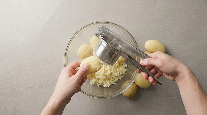 Hände pressen gekochte Kartoffel durch eine Kartoffelpresse in eine Glasschüssel mit feinem Kartoffelmus; mehrere geschälte Kartoffeln liegen daneben.