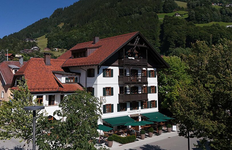 Ein traditionelles Hotel mit einem hellen, weiß gestrichenen Gebäude und roten Ziegeldächern. Es ist von Bäumen und einer schönen Landschaft umgeben, die Hügel und Wälder umfasst. Auf der Terrasse des Hotels stehen grüne Sonnenschirme und Tische für Gäste.