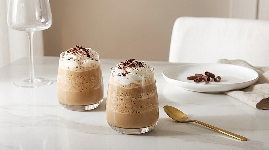 Zwei cremige Eiskaffees in kurzen Gläsern, jeweils mit Schlagsahne und Schokoraspeln, auf heller Marmoroberfläche, daneben goldener Löffel und kleiner Teller mit Schokoladenraspeln.