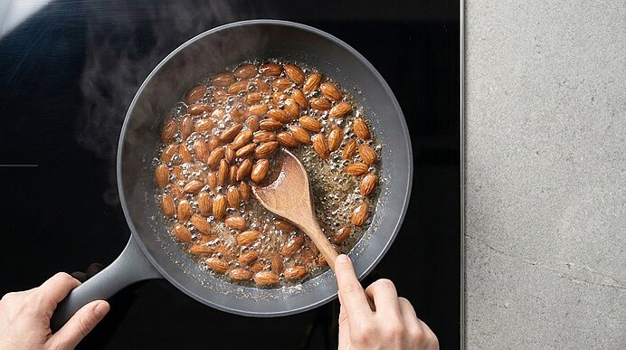 Hände rühren mit Holzlöffel ganze Mandeln in einer Pfanne, während sie auf dem Herd in Karamellsirup karamellisieren.