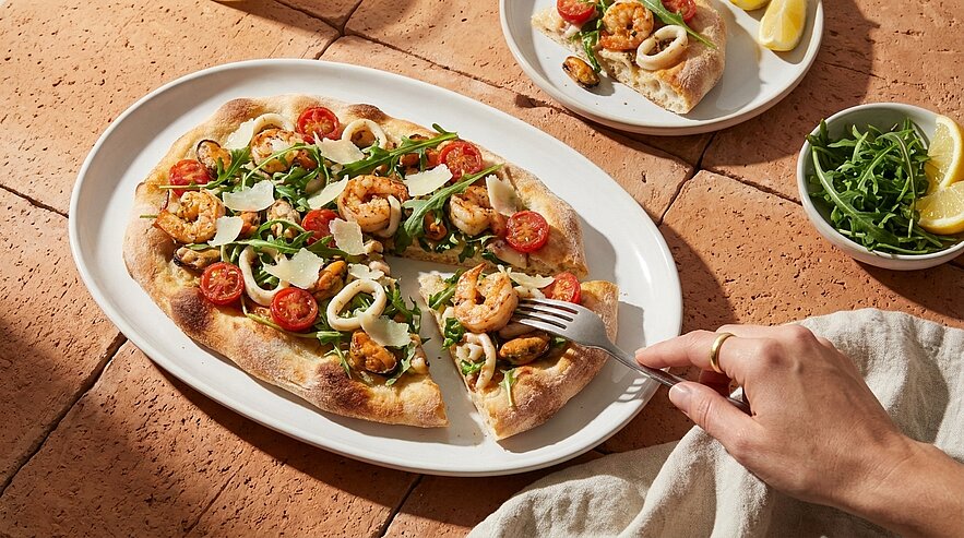 Eine handgehaltene Gabel schneidet ein Stück Pizza auf einem weißen Teller. Die Pizza ist belegt mit Garnelen, Tomaten, Zwiebeln und Rucola. Im Hintergrund sind weitere Teller mit Zitronenscheiben und Rucolasalat sichtbar. Der Untergrund ist aus terrakottafarbenen Fliesen.