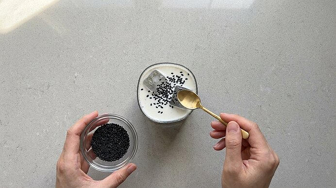 Hände von oben: Linke Hand hält eine kleine Glasschale mit schwarzen Sesamsamen, rechte Hand führt mit einem goldenen Löffel Samen in ein Glas mit milchigem Joghurt.