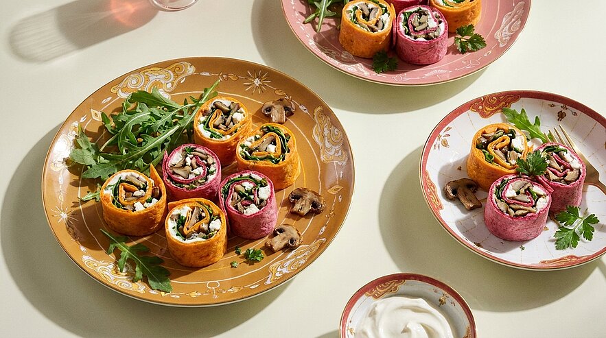 Mehrere bunte Wrap‑Rollen (pink und orange) gefüllt mit Gemüse auf Tellern, garniert mit Rucola, Champignons und Petersilie; kleine Schale mit weißem Dip.