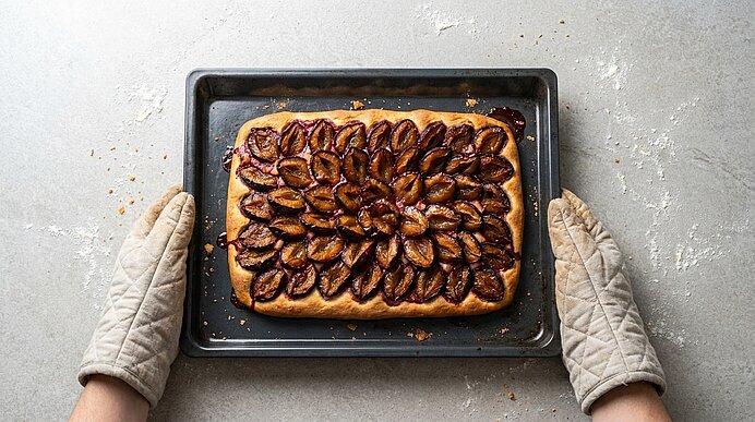 Frisch gebackener rechteckiger Pflaumenkuchen auf einem Backblech; dicht mit halbierten, glänzenden Pflaumen belegt. Zwei Hände mit Topfhandschuhen halten das Blech.