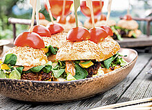 Eine rustikale Schale enthält mehrere kleine Burger, belegt mit frischem Gemüse. Jeder Burger ist mit einem Zahnstocher und einer Kirschtomate dekoriert. Die Umgebung wirkt einladend und natürlich, ideal für ein Buffet oder eine Feier im Freien.