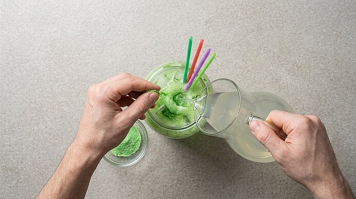 Zwei Hände gießen Zitronenlimonade in ein Glas mit grünem Drink und streuen Waldmeister-Brausepulver darüber.
