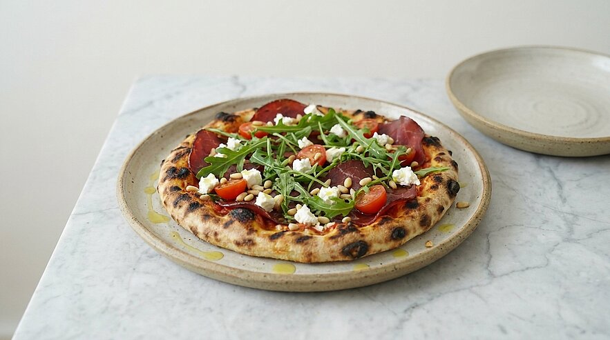 Pizza mit knuspriger, leicht gebräunter Kruste, belegt mit Rucola, halbierten Cherrytomaten, dünnen Schinken- oder Salami-Scheiben und kleinen Mozzarellastücken auf einem Teller.