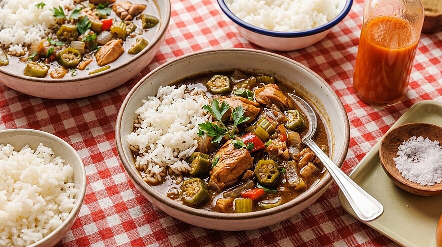 Schüssel mit Reis und würzigem Okra‑Hähnchen‑Eintopf, mit Petersilie garniert; daneben zusätzliche Reisschalen und eine Flasche scharfe Sauce.