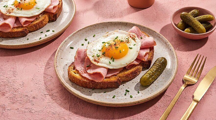 Offenes Sandwich: geröstetes Brot mit Schinken, Spiegelei mit weichem Dotter und Schnittlauch bestreut, dazu Gewürzgurke auf einem Teller.