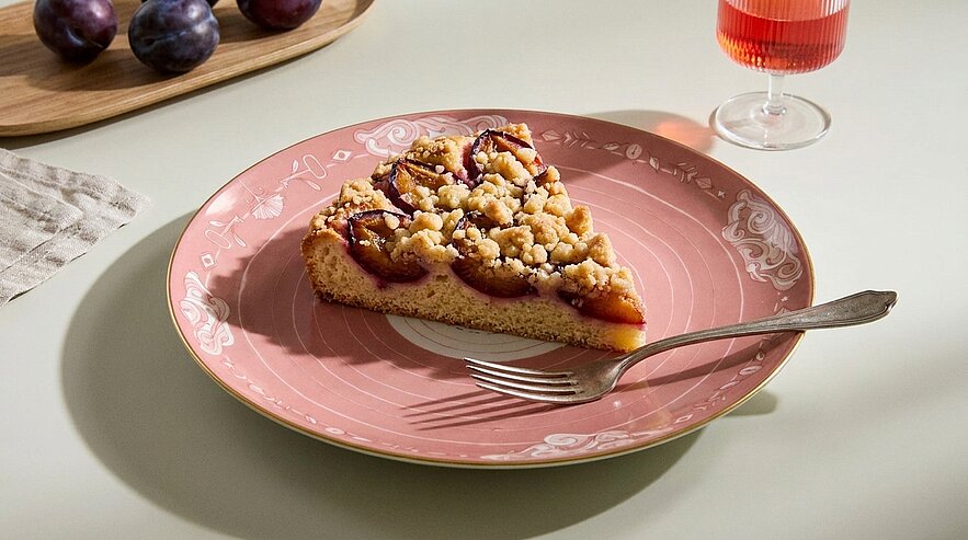 Dreieckiges Stück Pflaumen-Streuselkuchen auf einem rosa, dekorativen Teller, mit einer Gabel daneben.