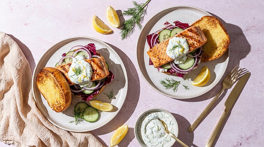 Zwei Teller mit gegrilltem Lachsfilet auf gerösteten Brötchenhälften, garniert mit Kräuter-Joghurtsoße, Gurkenscheiben, roten Zwiebeln, Dill und Zitronenspalten; daneben eine kleine Schüssel mit zusätzlicher Kräutersoße.