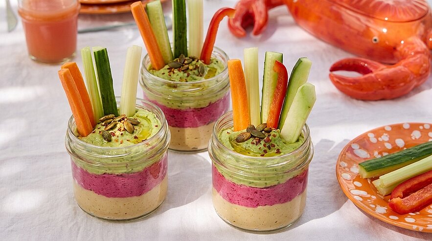 Drei kleine Gläser mit dreifarbigen Dips (beige, rosa, grün), bestreut mit Kürbiskernen und Gewürzen, darin Gemüsesticks (Karotte, Sellerie, Gurke, Paprika) stehend.