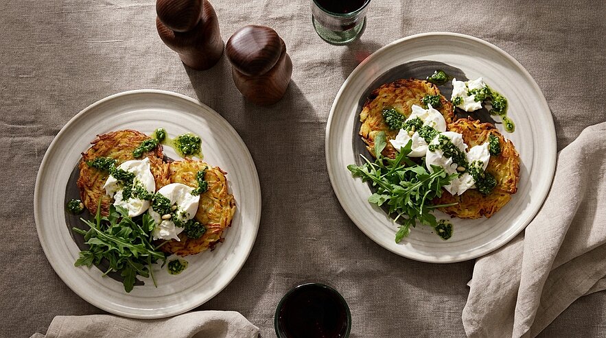 Zwei Teller mit knusprigen Kartoffelpuffern, darauf zerzupfte Burrata/Frischkäse, Rucola und grünes Kräuterpesto.