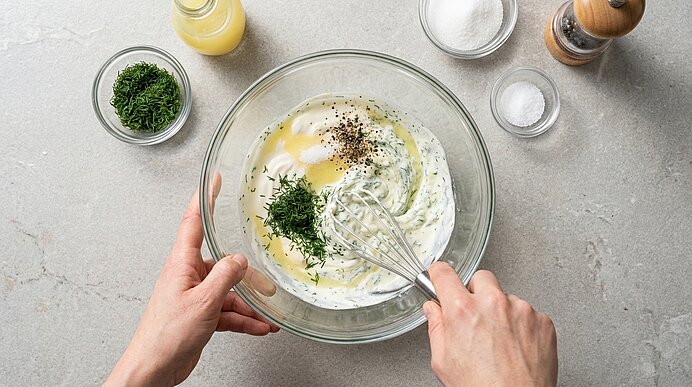 Zwei Hände rühren mit einem Schneebesen in einer Glasschüssel eine cremige Kräuter‑Dressing- oder Dipmischung mit Dill, Öl und Gewürzen.