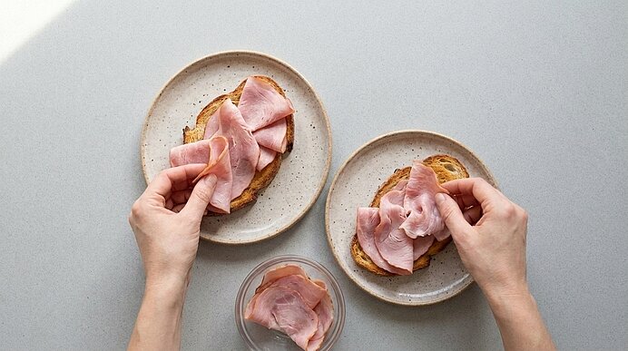 Hände belegen zwei Toasts auf Tellern mit dünnen Schinkenscheiben; daneben eine kleine Schüssel mit zusätzlichem Schinken.