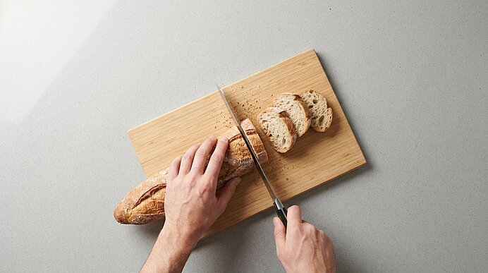Zwei Hände schneiden auf einem Holzbrett ein Baguette mit einem Kochmesser in Scheiben.