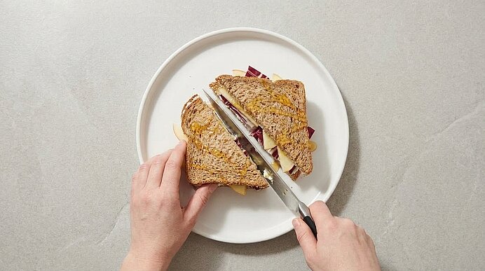 Zwei Hände halbieren eine Ziegenkäse-Stulle mit Apfel und Radicchio diagonal mit einem Messer auf einem Teller.