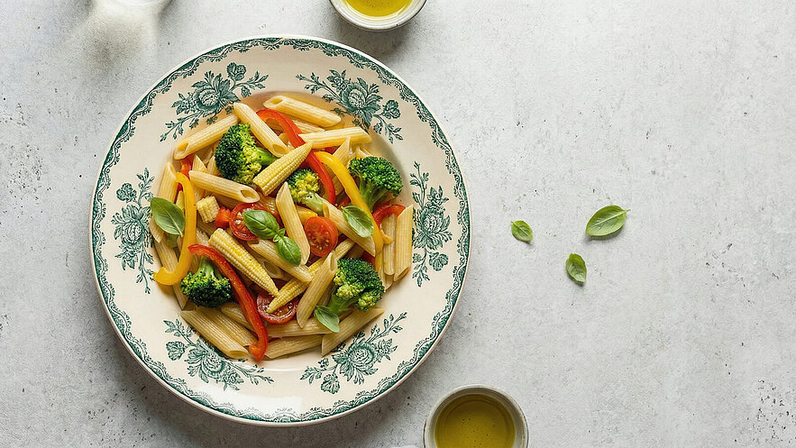 Eine Schale mit Penne-Nudeln, gemischt mit Brokkoli, Paprika, Cherrytomaten und Baby-Mais. Frische Basilikumblätter sind dekorativ hinzugefügt. Neben der Schale befindet sich ein kleiner Behälter mit Olivenöl. Der Hintergrund ist hell und neutral.