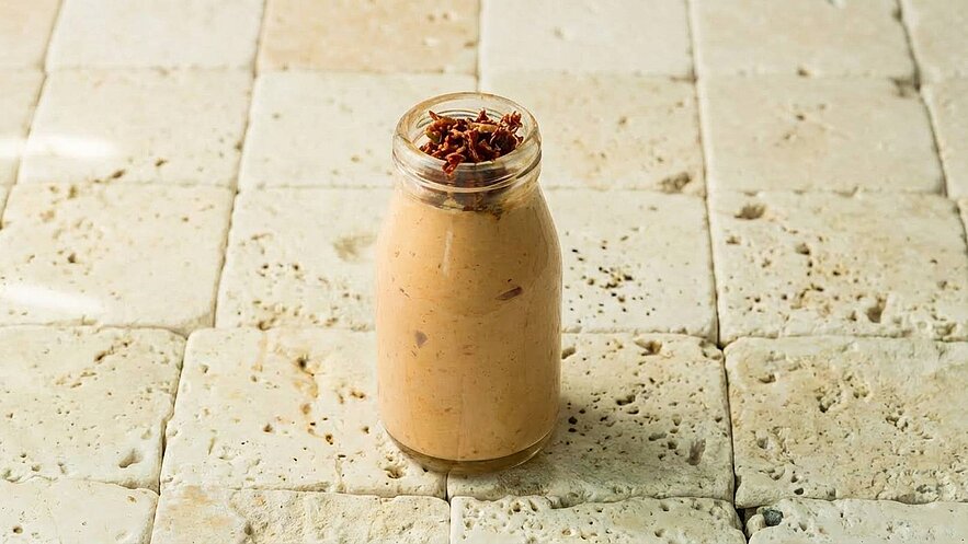 Ein Glas mit einer cremigen, beige-farbenen Paste ist auf einem strukturierten, hellen Untergrund platziert. Die Paste ist mit einer Schicht von knusprigen, braunen Stückchen oben dekoriert, die dekorativ wirken.