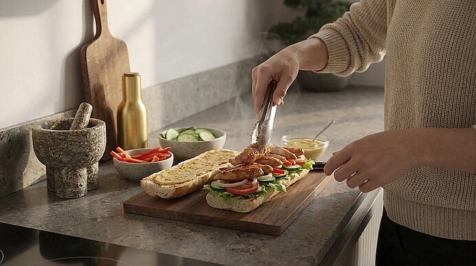 Person bereitet Sandwiches zu: Mit Zange legt sie heißes, gebratenes Fleisch auf Baguettehälften, belegt mit Tomaten-, Gurken- und Salatblättern auf einem Holzbrett.