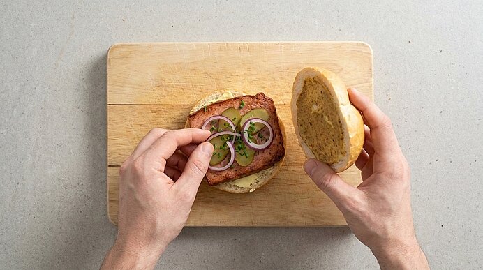 Zwei Hände belegen gebratenen Fleischkäse in einer Semmel mit Zwiebelringen und klappen die obere Hälfte darauf.