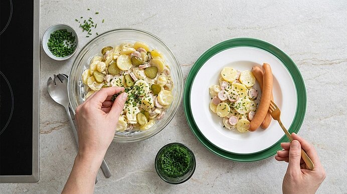 Hände servieren Kartoffelsalat mit Gewürzgurken und in Scheiben geschnittenen Würstchen; eine Hand streut Schnittlauch darüber.