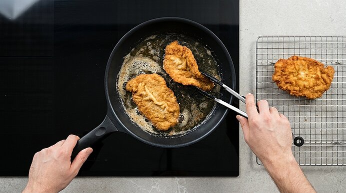 Zwei Hände wenden Schnitzel in heißem Butterschmalz in der Pfanne, während ein Schnitzel auf einem Gitter abtropft.