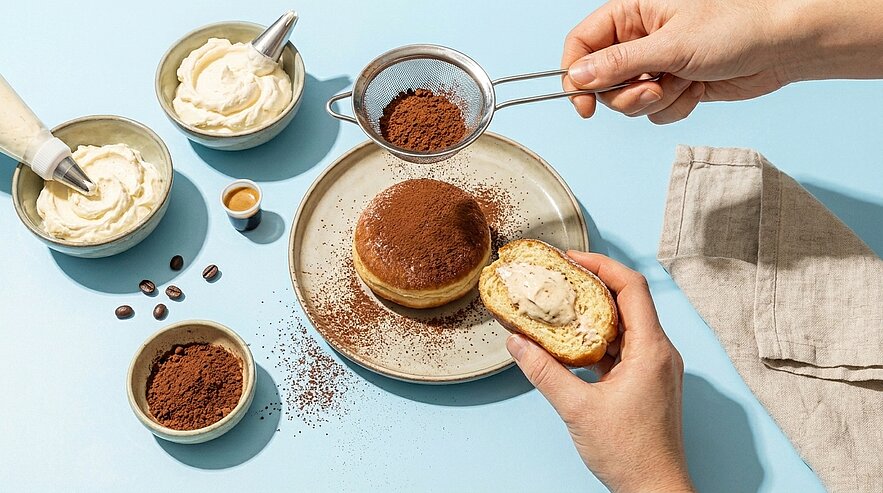 Hände bestäuben einen mit Creme gefüllten Krapfen auf einem Teller mit Kakao durch ein Sieb; eine Hand hält die aufgeschnittene Hälfte mit sichtbarer Füllung, Schalen mit Creme und Kakaopulver daneben.
