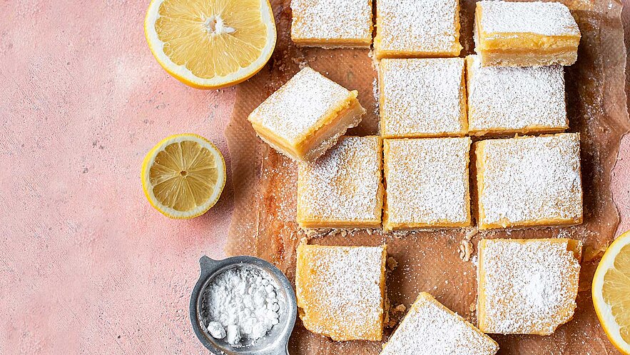Ein Tablett mit frisch gebackenem Zitronenkuchen, in gleichmäßige Stücke geschnitten und mit Puderzucker bestäubt. Neben dem Kuchen liegen zwei halbierte Zitronen. Ein kleines Schälchen mit Puderzucker befindet sich ebenfalls auf dem Tablett. Der Hintergrund ist blassrosa.
