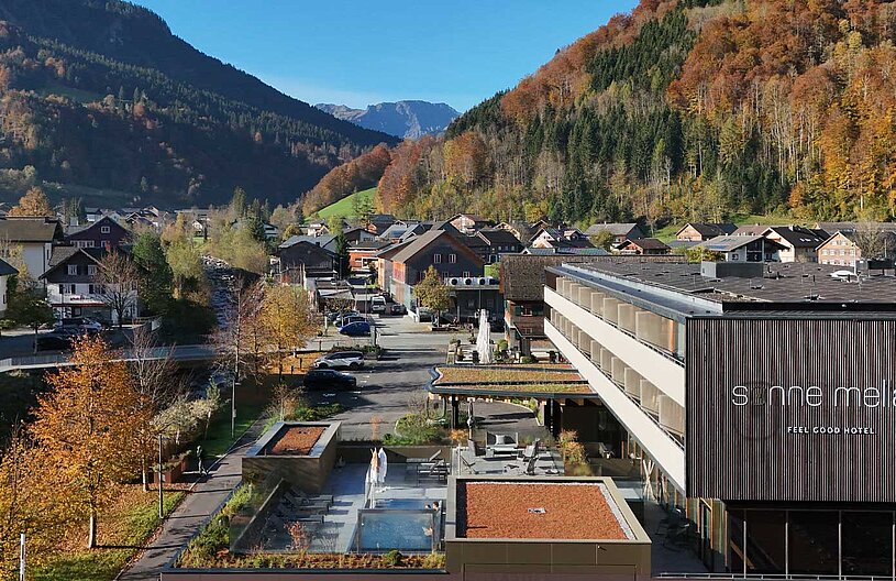 Das Bild zeigt die Umgebung des "Sonne Melau"-Hotels in den Bergen, umgeben von buntem Herbstlaub. Im Vordergrund sind moderne Gebäude und ein Außenbereich mit Sitzgelegenheiten und einem kleinen Pool zu sehen. Die Kulisse wird von sanften Hügeln und bewaldeten Bergen geprägt.