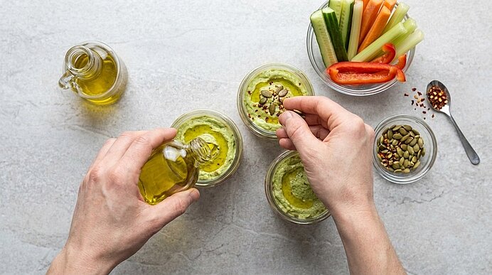 Zwei Hände garnieren Hummus in Gläsern mit Olivenöl und Kürbiskernen; daneben stehen Gemüsesticks.