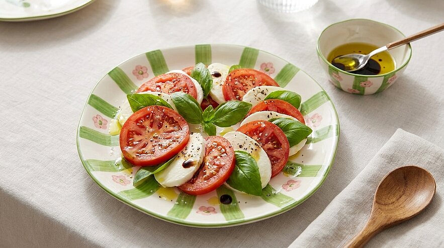 Caprese-Salat: abwechselnde Scheiben von Tomaten und Mozzarella, garniert mit frischen Basilikumblättern, Olivenöl, Balsamico sowie Salz und Pfeffer.
