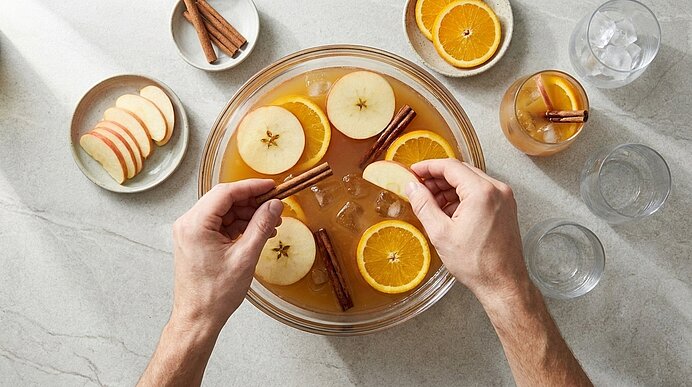 Zwei Hände legen eine Apfelscheibe in eine große Glasbowle mit Getränk, Orangenscheiben und Zimtstangen — Zubereitung eines gewürzten Herbst- oder Winterpunschs.