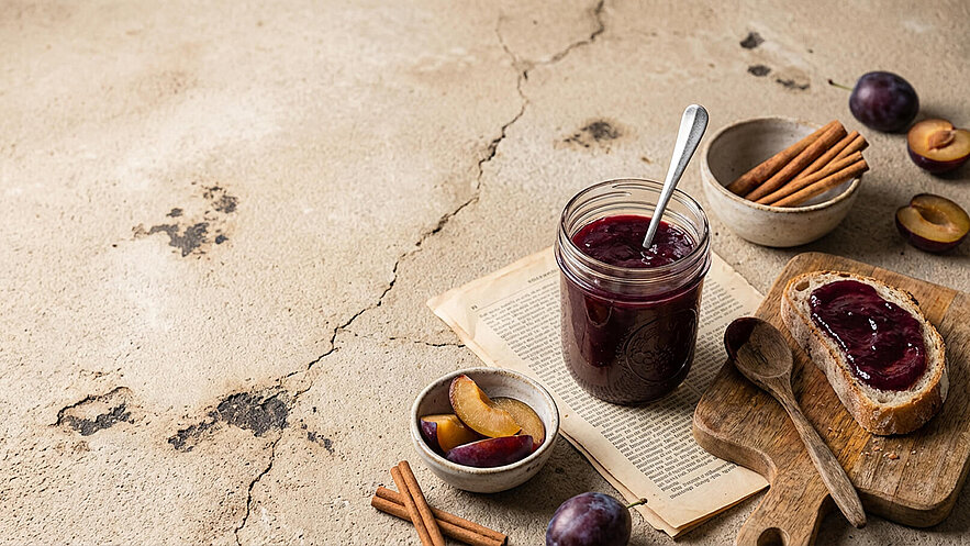 Ein Glas Pflaumenmarmelade mit einem Löffel steht auf einem Holzbrett neben einem belegten Brot. Daneben liegen frische Pflaumen, einige aufgeschnitten, sowie Zimtstangen in einer kleinen Schüssel. Die Hintergrundfläche ist rau und unregelmäßig.