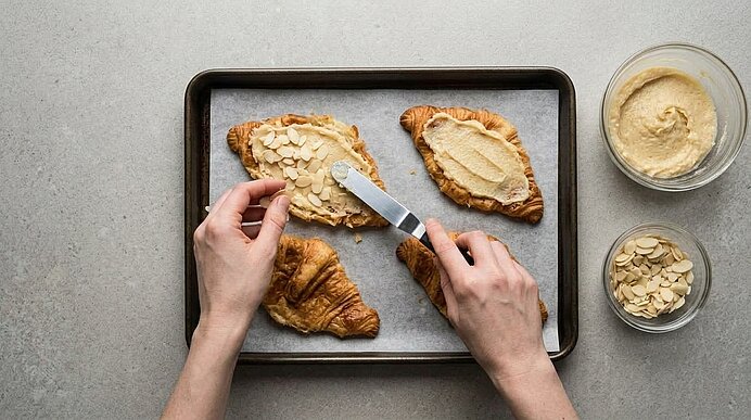 Zwei Hände streichen Mandelcreme auf flache Croissants auf einem Backblech und streuen Mandelblättchen darüber.