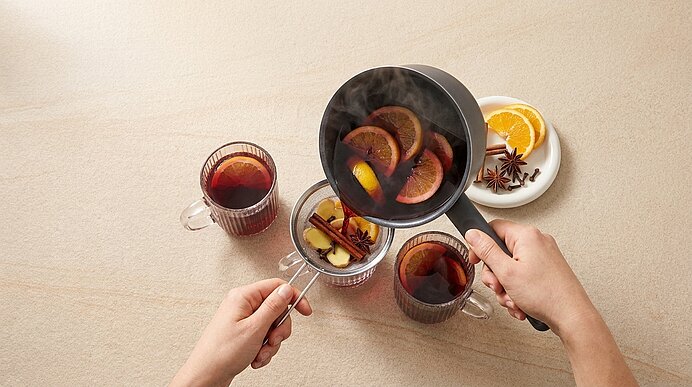 Zwei Hände seihen dampfenden Glühwein mit Orangen- und Gewürzstücken aus einem kleinen Topf durch ein Sieb in transparente Tassen; Teller mit Orangen, Sternanis und Zimt daneben.