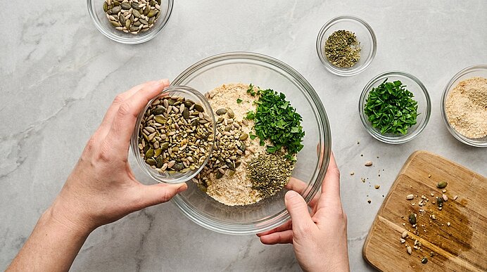 Zwei Hände geben Kürbiskerne in eine gläserne Schüssel mit Semmelbröseln, gehackter Petersilie und Gewürzen zum Vermengen.