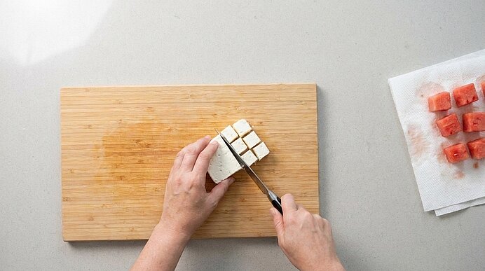 Zwei Hände schneiden Feta auf einem Holzbrett in Würfel. Daneben liegen Wassermelonenwürfel auf Küchenpapier.