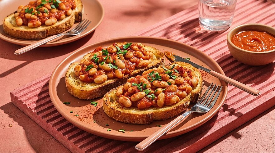Zwei Scheiben geröstetes Brot mit warmen weißen Bohnen in Tomatensauce, mit gehackter Petersilie bestreut, serviert auf einem Teller; daneben eine kleine Schale mit zusätzlicher Sauce.